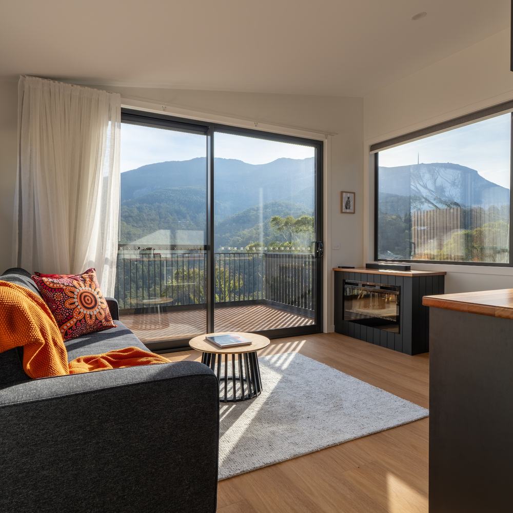 Living room of ironbark cabin with lounge, fireplace, kitchen looking out to kunanyi view