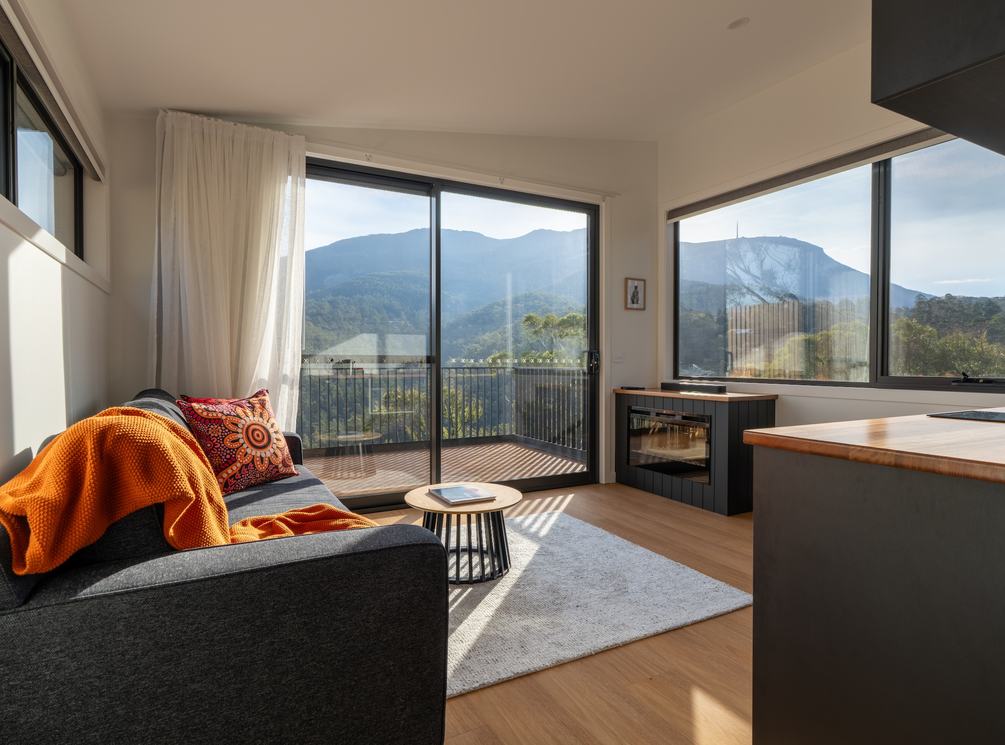 Living room of ironbark cabin with lounge, fireplace, kitchen looking out to kunanyi view