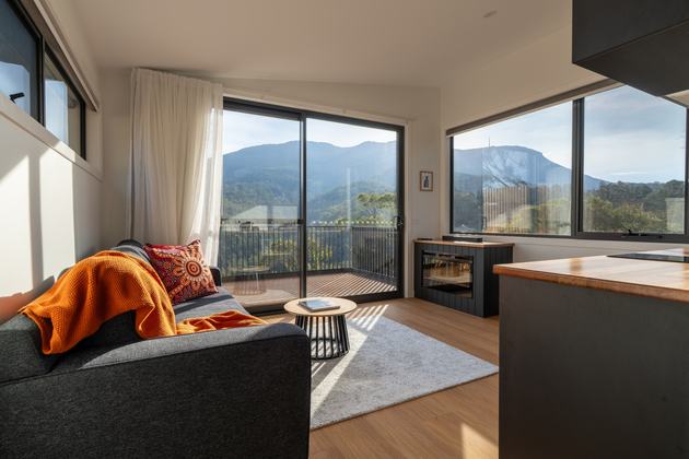 Living room of ironbark cabin with lounge, fireplace, kitchen looking out to kunanyi view